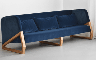 A modern sofa featuring deep blue fabric upholstery and a distinctive exposed light-toned wooden frame. The sofa has a single seat cushion and three loose back cushions, with a gently sloped back and integrated armrests that flow into the overall form. The unique wooden frame forms an angular support structure beneath the upholstered body. — 3D model