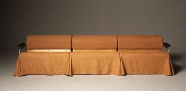 A long, rectangular sofa with a curved bolster backrest. The sofa is upholstered in a textured, light brown fabric. It features a metal frame with armrests that have black accents. The base of the sofa is covered by a draped skirt of the same fabric, creating a segmented appearance. — 3D model