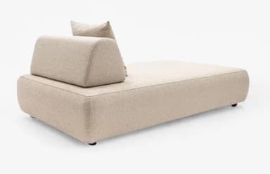 A modern daybed featuring a minimalist design. It is upholstered in a light beige, textured boucle fabric, with distinct piping along the edges. The base is long and rectangular with softly rounded corners. At one end, there is a large, loose back cushion and a smaller, square throw pillow, both matching the main upholstery. The daybed sits on small, discreet dark feet. — 3D model
