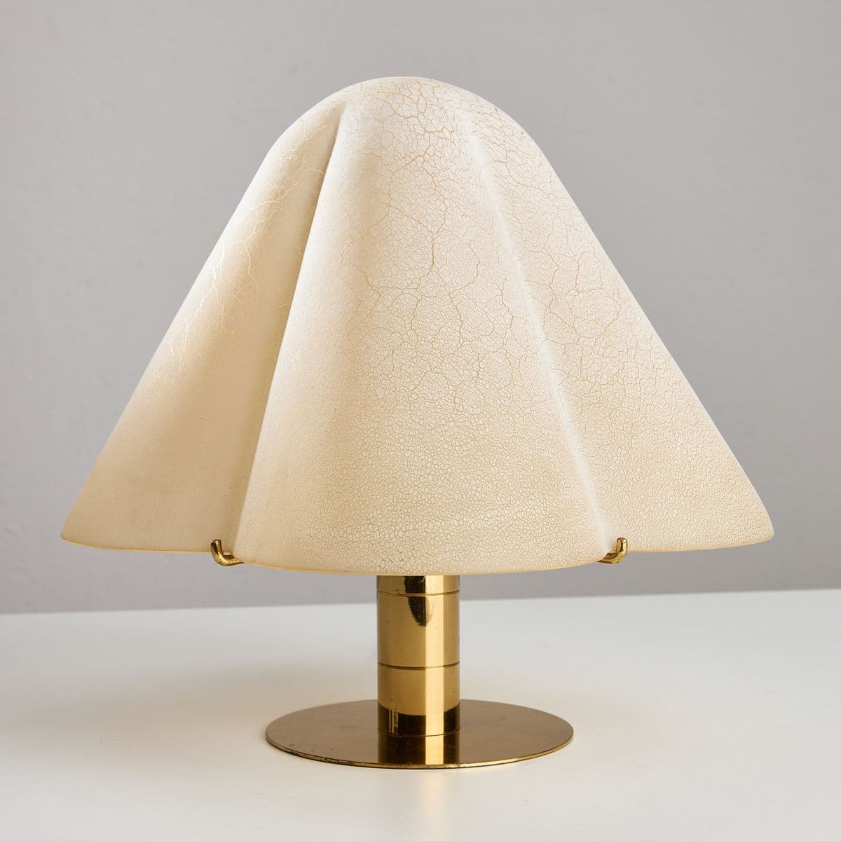 A table lamp with a unique, draped lampshade. The lampshade is a pale cream color with a crackled finish that creates a textured surface. It flares out from a central support and is held in place by small brass clips. The base and central stem are made of polished brass, with the stem featuring stacked cylindrical elements. The overall design is modern and sculptural. — 3D model