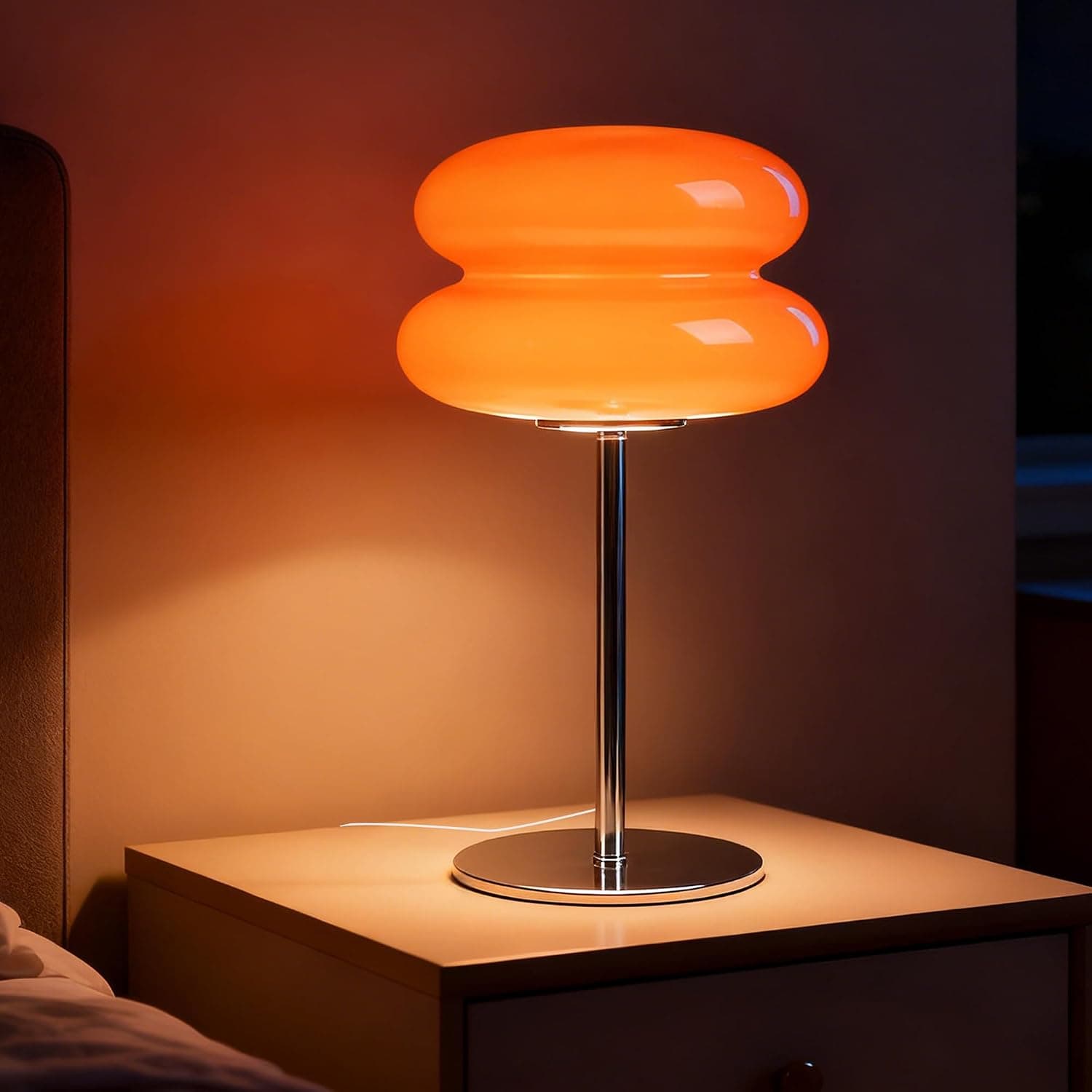 An illuminated table lamp featuring an orange, hourglass-shaped glass shade composed of two stacked, rounded sections. The shade is supported by a slender chrome-finished metal stem that rises from a flat, circular chrome base. The lamp emits a warm, diffused orange glow, indicative of its material and the light source within. — 3D model