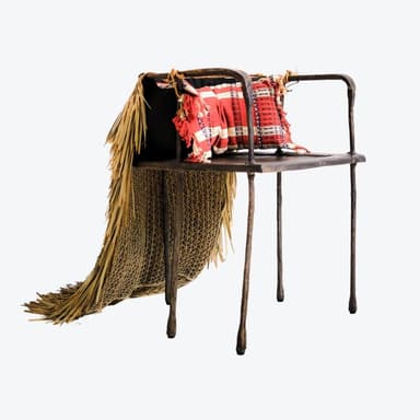 A side table with a dark, metallic frame featuring two thin legs on the visible side. The tabletop is obscured by a long, draped skirt made of dried grass or palm fronds. The grass material is layered and reaches the floor, creating a voluminous effect. There are visible red beads and rope details on the frame near the top. — 3D model
