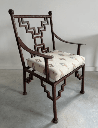 An armchair with a dark brown, possibly metal, frame featuring a geometric, stepped pattern on the back and legs. It has curved armrests and a cushioned seat upholstered in a textured fabric with a geometric pattern in shades of beige, brown, and grey. The chair has a decorative finial at the top of each back leg and round feet. — 3D model