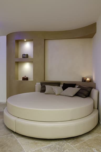 A large, round upholstered daybed featuring a two-tiered design with a main circular seating platform and a lower, wider circular base. It includes a partial backrest and is accessorized with multiple throw pillows. — 3D model