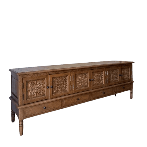 A long, rectangular wooden sideboard with six doors and three drawers. The doors feature intricate carved floral and geometric patterns. The drawers are positioned below the doors and have small, round dark knobs. The piece stands on four turned legs, two visible at the front and two at the rear, with a plinth base connecting the lower section. — 3D model