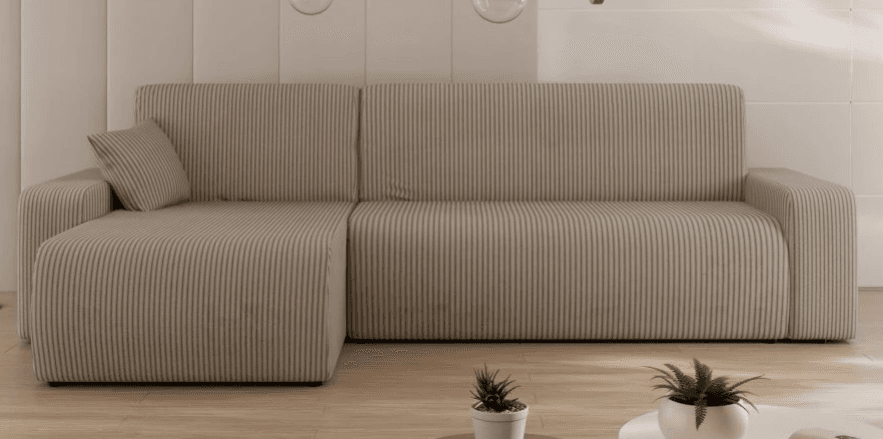 A modular sectional sofa with a chaise lounge extension. The sofa features a ribbed upholstery texture and rounded edges on its frame and armrests. A single matching throw pillow is visible on the chaise section. The sofa is upholstered in a light brown or beige fabric. — 3D model