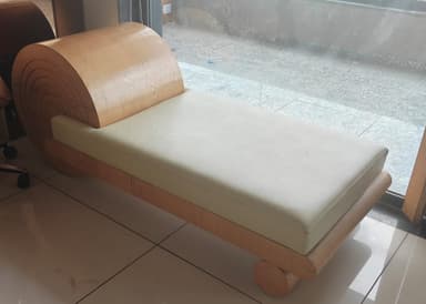 A modern chaise lounge or daybed featuring a minimalist design with a light-toned wooden frame and a cream-colored upholstered cushion. The most striking feature is the large, cylindrical rolled headrest made from bent wood, displaying concentric grain patterns on its ends. The base consists of a low-profile rectangular frame supported by integrated cylindrical wooden legs. — 3D model