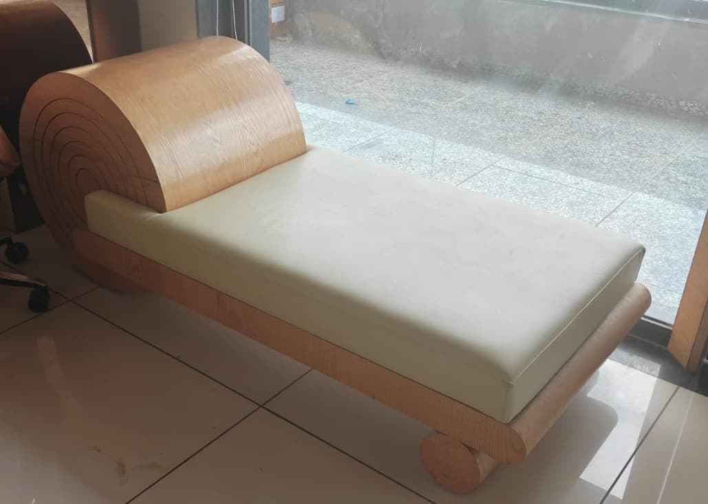 A modern chaise lounge or daybed featuring a minimalist design with a light-toned wooden frame and a cream-colored upholstered cushion. The most striking feature is the large, cylindrical rolled headrest made from bent wood, displaying concentric grain patterns on its ends. The base consists of a low-profile rectangular frame supported by integrated cylindrical wooden legs. — 3D model