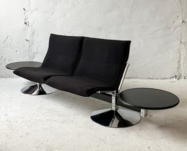 A two-seater sofa with attached side tables. The sofa features black upholstered seats and backrests with a ribbed texture on the backrests. The frame appears to be metal, possibly chrome or stainless steel, with a dark finish on the main horizontal support. Each seat has a cantilevered base with a circular, flared chrome pedestal. Attached to the horizontal frame on either side are circular, dark-colored side tables with a glossy finish, supported by metal brackets. — 3D model