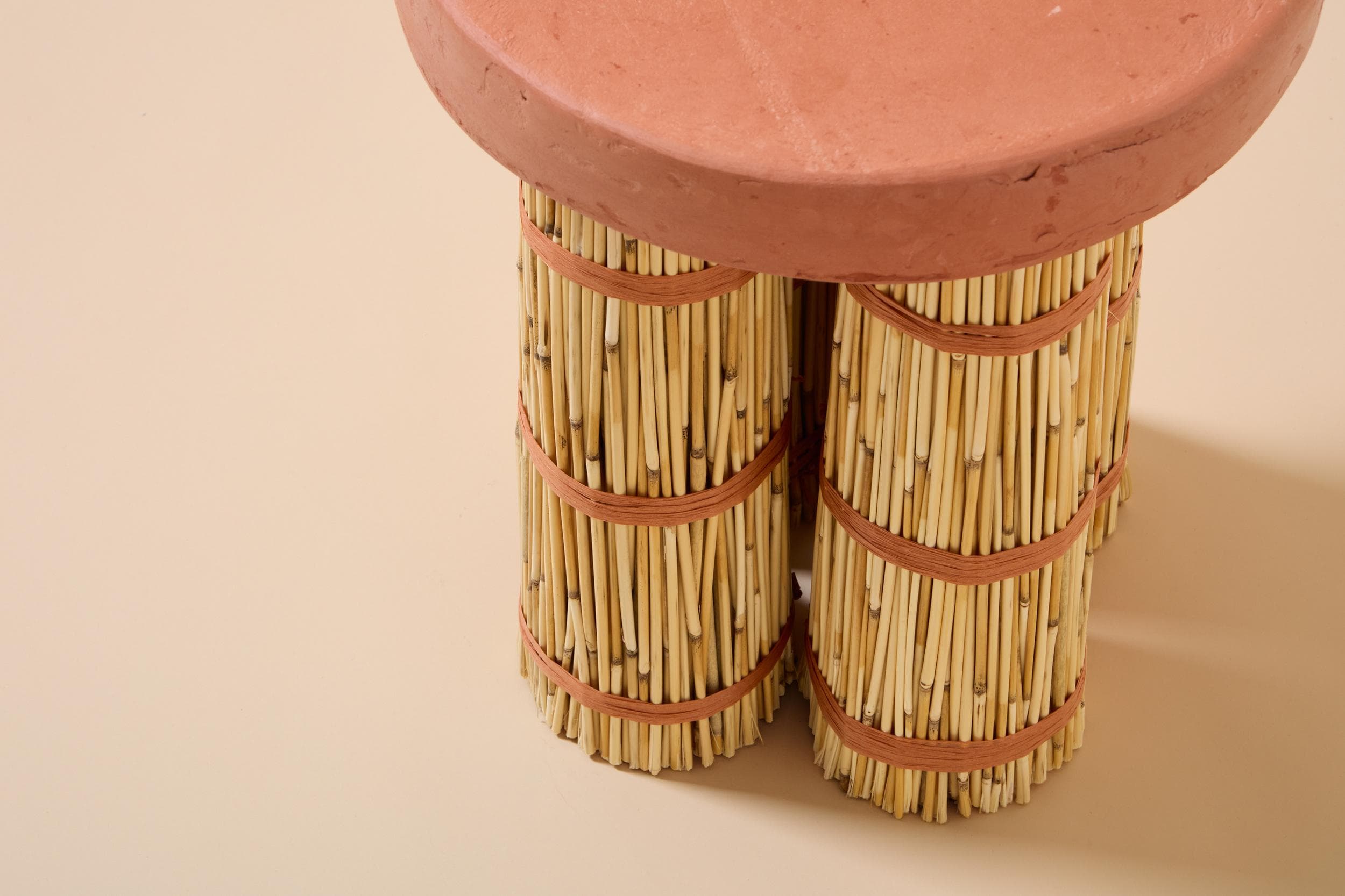 A small, round accent table featuring a flat, circular top made of reddish-brown terracotta. The base consists of three bundles of light-colored bamboo sticks, tied securely in multiple places with reddish-brown woven natural fiber. The legs appear to be cylindrical and provide a sturdy base for the rustic top. — 3D model