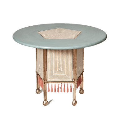 A decorative round accent table featuring a pale blue-green top with a central pentagonal geometric inlay. The base is pentagonal, constructed with a cream-colored material featuring intricate geometric patterns, framed by a gold-toned metallic structure. A fringe of coral pink tassels hangs from the lower edge of the base. The table stands on four short, ornate gold-toned metallic legs with ball feet. — 3D model