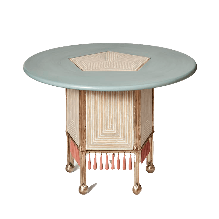 A decorative round accent table featuring a pale blue-green top with a central pentagonal geometric inlay. The base is pentagonal, constructed with a cream-colored material featuring intricate geometric patterns, framed by a gold-toned metallic structure. A fringe of coral pink tassels hangs from the lower edge of the base. The table stands on four short, ornate gold-toned metallic legs with ball feet. — 3D model