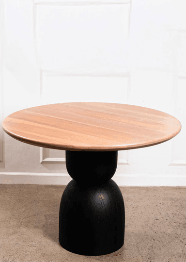 A contemporary round side table featuring a light-toned wooden top and a matte black sculptural pedestal base. The base has a distinctive hourglass-like shape with subtle vertical lines or grooves, giving it a tactile texture and modern aesthetic. The table presents a two-tone appearance, blending natural wood with a bold black finish. — 3D model