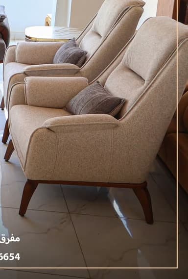 A modern upholstered armchair featuring a beige textured fabric, a high back, and integrated armrests. It is supported by four dark brown tapered wooden legs. A small rectangular grey cushion is positioned on the seat back for additional lumbar support. — 3D model