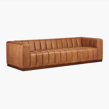 A long, channel-tufted sofa with a minimalist design. The sofa features a continuous padded back and armrest with vertical channels, upholstered in a tan leather-like material. It rests on a low, dark wood plinth base. The overall aesthetic is modern with a touch of art deco. — 3D model