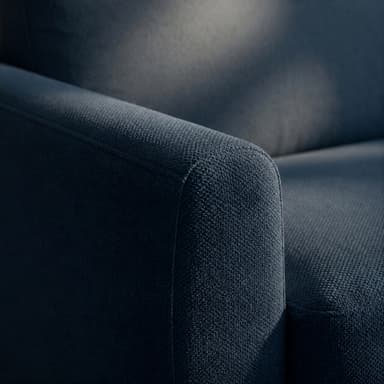 A close-up view of the armrest and cushions of a contemporary dark blue upholstered sofa. The sofa features clean lines, fixed rectangular armrests with a slight outward curve, and plump, separate seat and back cushions, all covered in a finely textured woven fabric. Small, dark, recessed feet are visible at the base. — 3D model