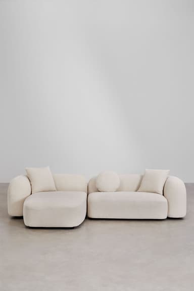 A contemporary L-shaped sectional sofa consisting of two modular pieces: a main sofa unit and an extended chaise lounge. The entire piece is upholstered in a textured, light-colored boucle fabric. It features a low-profile design with rounded edges and soft, voluminous forms, giving it a plush appearance. The sofa includes three accent pillows: two square and one cylindrical bolster pillow, all matching the primary upholstery. The design emphasizes comfort and a minimalist, inviting aesthetic. — 3D model