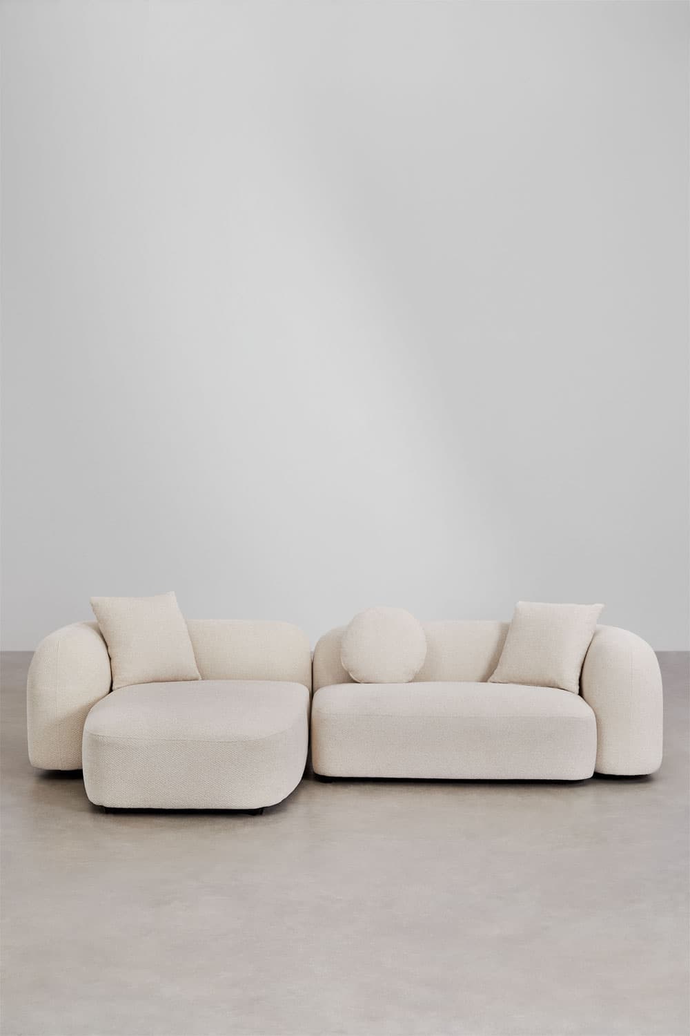A contemporary L-shaped sectional sofa consisting of two modular pieces: a main sofa unit and an extended chaise lounge. The entire piece is upholstered in a textured, light-colored boucle fabric. It features a low-profile design with rounded edges and soft, voluminous forms, giving it a plush appearance. The sofa includes three accent pillows: two square and one cylindrical bolster pillow, all matching the primary upholstery. The design emphasizes comfort and a minimalist, inviting aesthetic. — 3D model