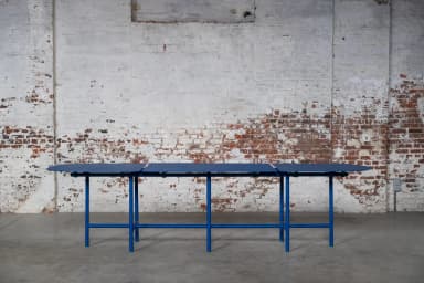 A long, modular dining table featuring three distinct segments with rounded ends, supported by a vibrant blue tubular metal frame. Each segment's top is dark blue, complementing the bright blue base, with small green connectors and feet accents. — 3D model