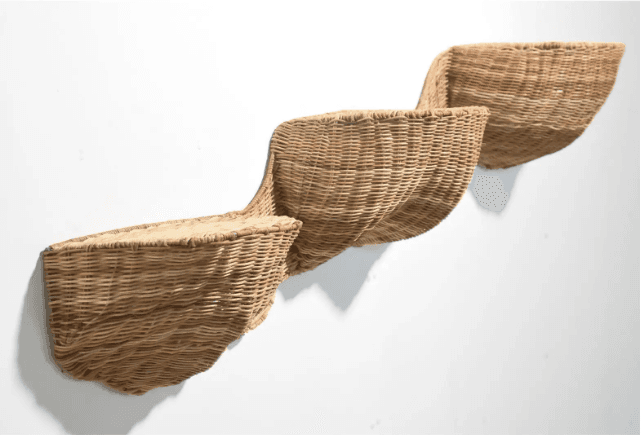 A trio of wall-mounted woven wicker seats arranged in a staggered, ascending pattern. Each seat is a distinct, curved basket-like form, constructed from natural-colored wicker. The design appears to be avant-garde, with the seats projecting outwards from a white wall. The closest seat on the left is the largest and lowest, with the subsequent seats progressively smaller and higher. — 3D model