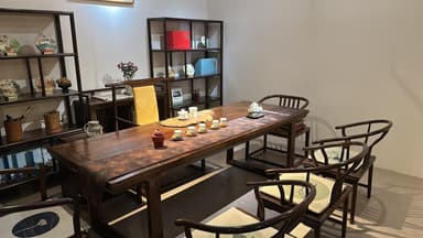 A dark brown rectangular solid wood table, likely used for dining or tea ceremonies. It features a thick, substantial tabletop with a rich, dark stained finish that reveals the natural wood grain. The table stands on two inverted U-shaped or Z-shaped legs, providing a stable and visually minimalist base. A patterned fabric runner with a traditional design is draped across the tabletop, adorned with a selection of tea ware and decorative items. — 3D model