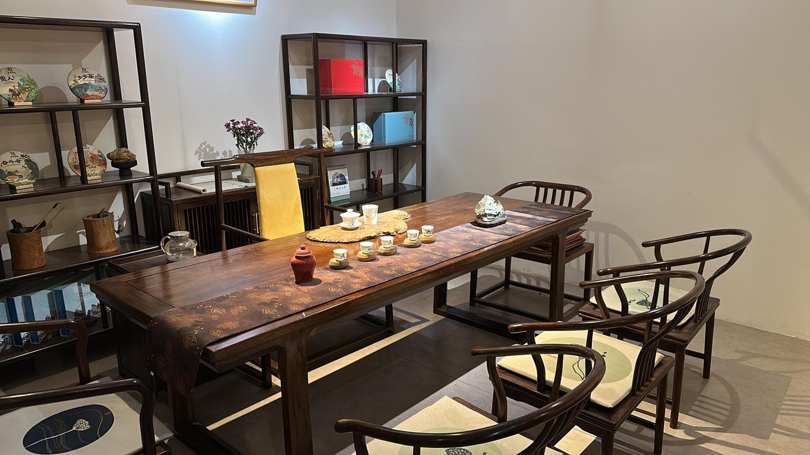 A dark brown rectangular solid wood table, likely used for dining or tea ceremonies. It features a thick, substantial tabletop with a rich, dark stained finish that reveals the natural wood grain. The table stands on two inverted U-shaped or Z-shaped legs, providing a stable and visually minimalist base. A patterned fabric runner with a traditional design is draped across the tabletop, adorned with a selection of tea ware and decorative items. — 3D model