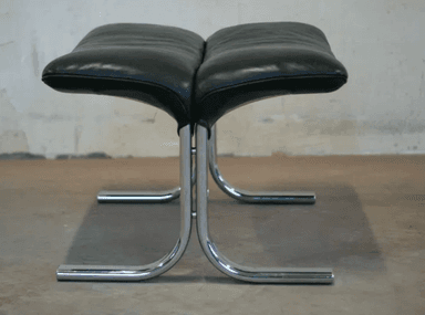A black leather armchair with a matching ottoman. The chair features a high back and a curved, cantilevered chrome base. The ottoman also has a curved chrome base and is upholstered in matching black leather with a segmented cushion design. — 3D model