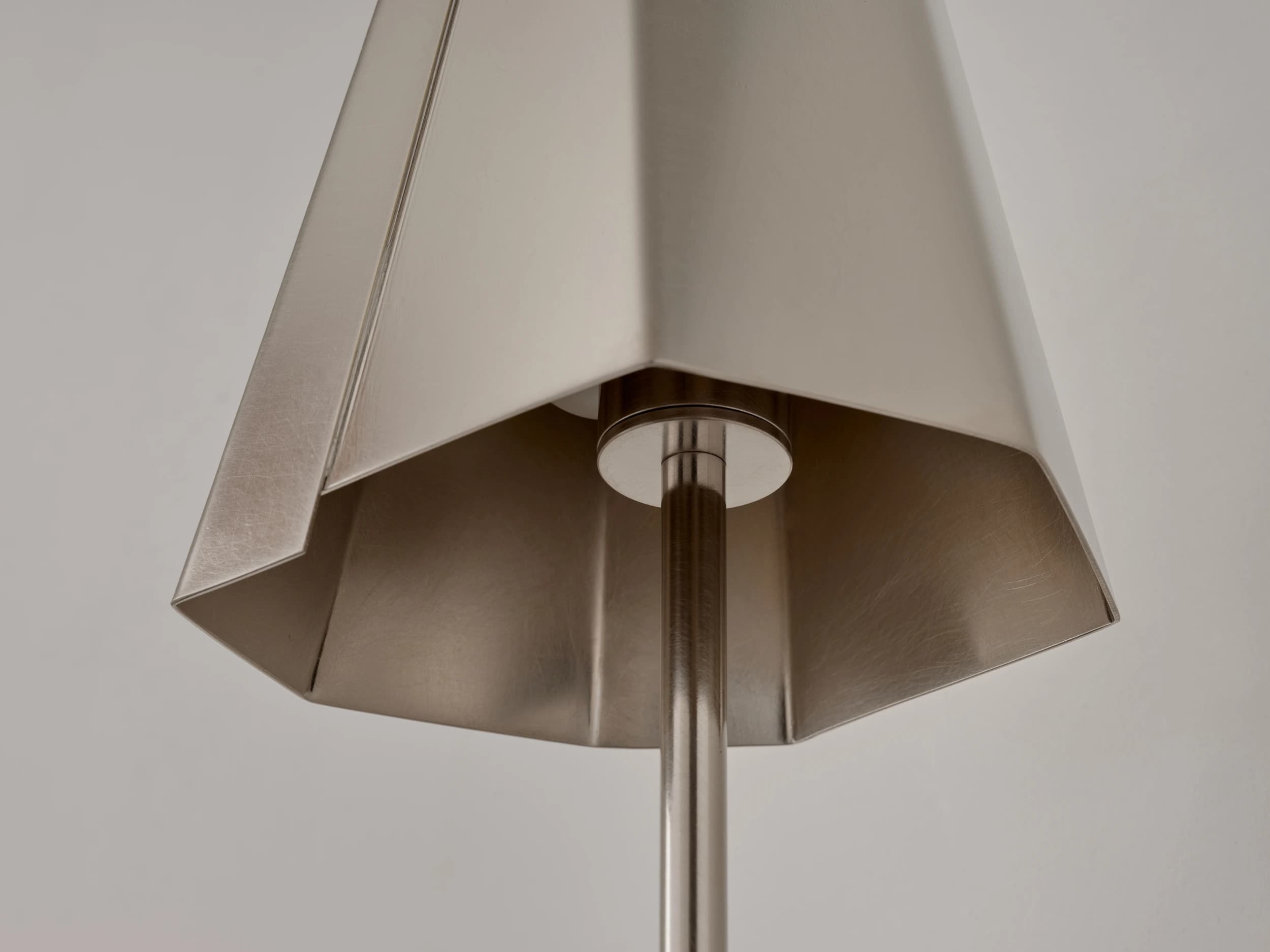 A close-up image of a modern pendant light's hexagonal, faceted shade, crafted from brushed metal. A central rod passes through the bottom opening, connecting to the light source or suspension mechanism. The shade features distinct angular folds, giving it a geometric appearance. — 3D model