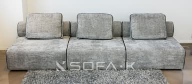 A contemporary, low-profile modular sofa upholstered in a textured, speckled grey fabric. The sofa consists of three distinct seating modules, each with a loose seat cushion and a loose rectangular back cushion. The back cushions feature black piping around their edges. The sofa integrates low, rounded or rolled armrests at each end, which blend seamlessly with the overall low-profile design. A dark line of piping or stitching runs along the seams of the modular units and along the bottom edge of the sofa, creating a clean, defined aesthetic. The sofa rests directly on the floor, lacking visible legs. — 3D model