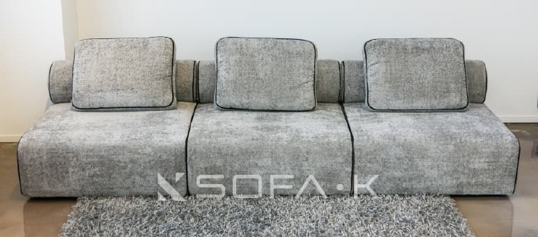 A contemporary, low-profile modular sofa upholstered in a textured, speckled grey fabric. The sofa consists of three distinct seating modules, each with a loose seat cushion and a loose rectangular back cushion. The back cushions feature black piping around their edges. The sofa integrates low, rounded or rolled armrests at each end, which blend seamlessly with the overall low-profile design. A dark line of piping or stitching runs along the seams of the modular units and along the bottom edge of the sofa, creating a clean, defined aesthetic. The sofa rests directly on the floor, lacking visible legs. — 3D model