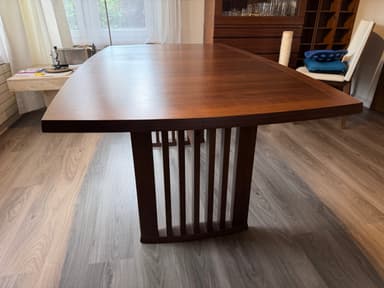 A modern dining table featuring a dark brown wooden top with elegantly curved edges and a matching slatted base. The table top displays a visible wood grain pattern, suggesting a veneer finish, and shows some minor surface wear. The base appears robust, designed with vertical wooden slats, which likely form a pedestal or trestle-style support. — 3D model