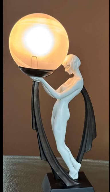 A striking Art Deco style figural table lamp featuring a nude female figure in a light finish, gracefully supporting a large, spherical frosted glass shade. The figure is integrated into a dark, flowing base and support structure with geometric accents, creating a high-contrast decorative lighting piece. — 3D model