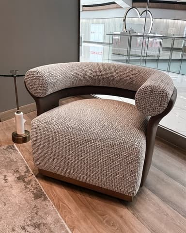A modern barrel-back armchair featuring a rounded, enveloping form. The main body is upholstered in a textured, light brown or beige fabric, complemented by smooth, dark brown leather or faux leather accents on the outer back and sides. The chair rests on a recessed, dark brown base, suggesting a low profile. — 3D model