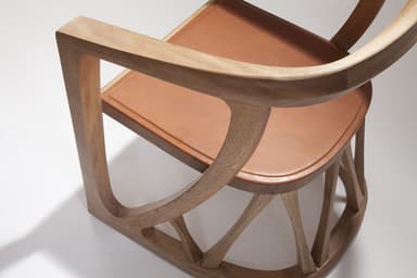A contemporary armchair crafted from light-colored solid wood, featuring a gracefully curved backrest and integrated armrests. The chair incorporates a geometric, sculptural base with multiple intersecting wooden struts providing support. A contrasting reddish-brown leather cushion forms the seat, complementing the natural wood tone. — 3D model