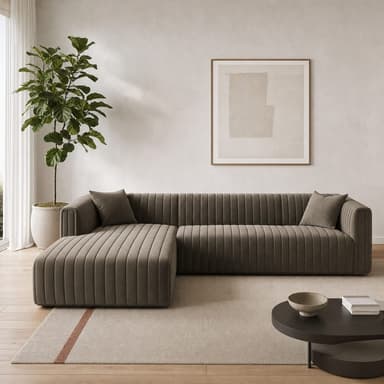 An L-shaped sectional sofa featuring plush channel-tufted upholstery in a neutral taupe or grey velvet fabric. The design is modern with clean lines, integrated armrests that flow into the backrest, and a distinct chaise lounge extension. Two matching square throw pillows are included. — 3D model