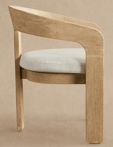 A contemporary dining armchair featuring a solid wood frame with a light, natural finish and visible grain. It has a distinctive curved backrest that seamlessly transitions into integrated armrests. The seat is upholstered with a light-colored, textured fabric cushion. The legs are sturdy and straight, giving the chair a minimalist yet robust appearance. — 3D model