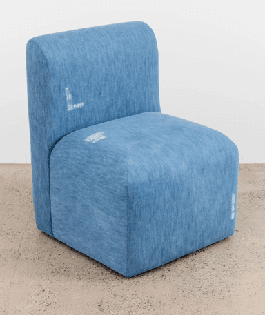 A modern armchair with a compact, blocky design. It features a low-profile seat and a slightly curved, integrated backrest. The upholstery is a textured, medium-blue fabric, possibly denim or a similar textile. Visible white graphic elements, resembling stylized text or symbols, are present on the backrest and seat. — 3D model
