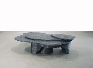 A sculptural, low-lying table featuring an organic, multi-tiered design. The dark, matte material has a layered texture, mimicking natural rock formations or topographical maps. The table top comprises several irregularly shaped, overlapping planes at varying heights, supported by an equally irregular and textured base. — 3D model