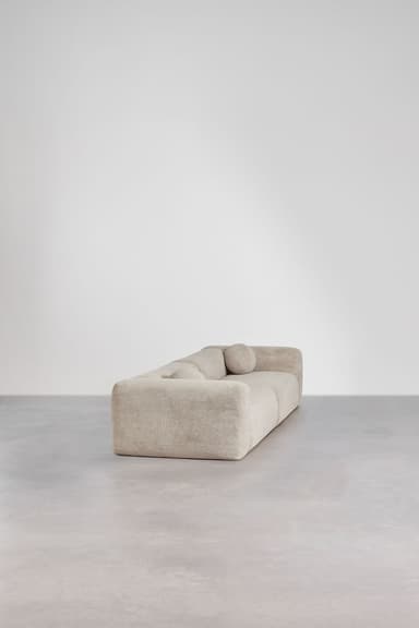 A long, low-profile modular sectional sofa upholstered in a light greige, textured fabric. The sofa consists of at least three distinct segments, featuring rounded edges and a minimalist, contemporary design. It sits directly on the floor with no visible legs, giving it a substantial and soft appearance. — 3D model