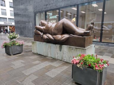 A large bronze sculpture depicting a reclining female figure, situated outdoors on a substantial concrete plinth in front of a modern glass building. The figure has soft, rounded forms and is posed with her head resting on a raised form resembling a pillow, with her body gently draped. — 3D model