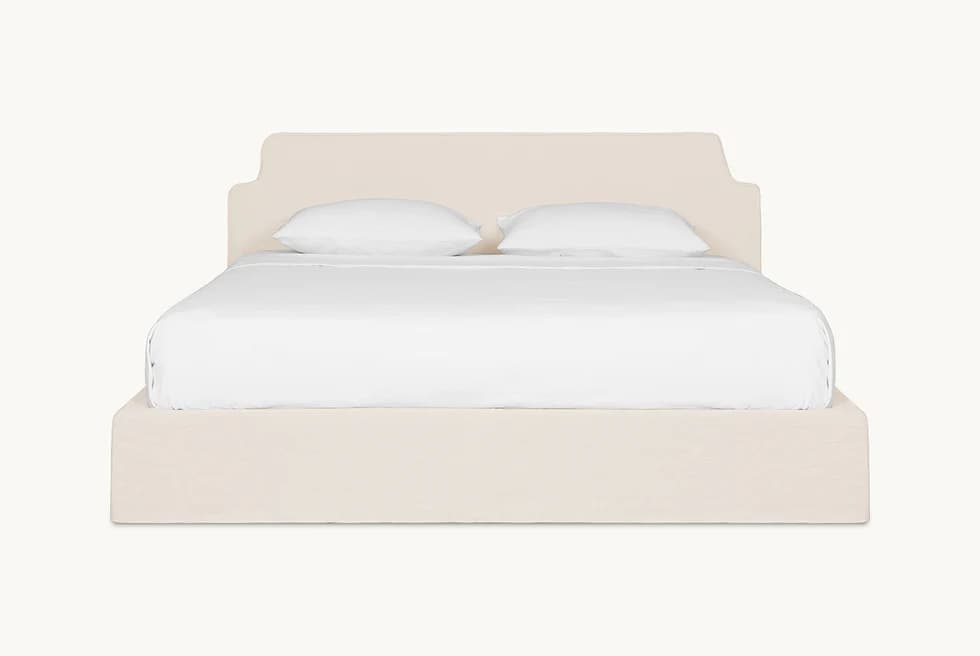 A bed frame with a padded headboard and platform base, covered in a neutral-toned fabric. The headboard has a distinctive silhouette with curved upper corners. The bed is shown with white linens and two pillows. — 3D model