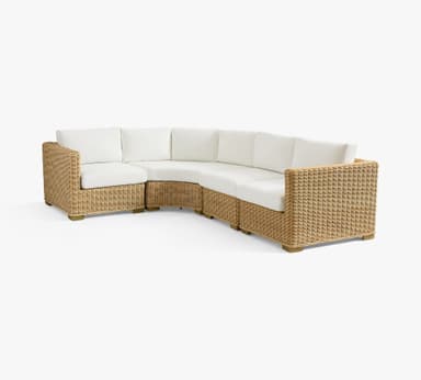 A modular sectional sofa with a curved design. The base and arms are constructed from woven wicker or rattan material, with a natural tan color. It features thick, white cushioned seating and backrests. The sofa is elevated on short, light-colored wooden legs. — 3D model