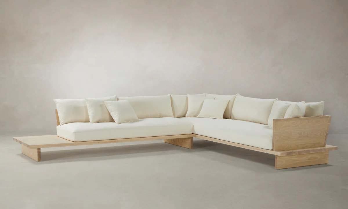 A modular sectional sofa with a low-profile design. The frame appears to be constructed from light-colored wood, possibly ash or oak, with visible grain. The sofa features generous, plush cushions upholstered in a cream or off-white fabric. It has a L-shaped configuration with an extended side surface on one end, functioning as a bench or table. The backrests are composed of multiple large cushions. — 3D model