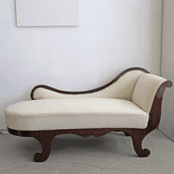 An elegant classical chaise lounge featuring a dark wood frame and light cream-colored upholstery. The frame exhibits distinctive scrolled and carved elements, particularly on the backrest/armrest and the base. The design suggests a refined, antique aesthetic, often associated with Regency or Empire styles. The upholstery is a plain, light fabric, providing a contrast to the dark wood. — 3D model