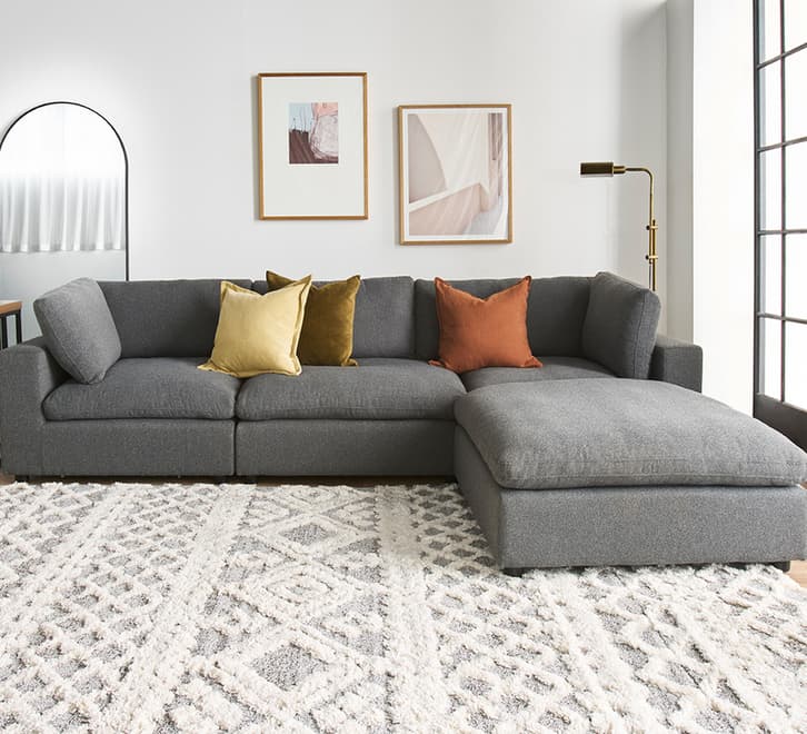 A contemporary gray fabric sectional sofa featuring a modular design, an L-shaped configuration, and a chaise lounge extension. It includes loose back cushions and is accessorized with several accent pillows in warm tones. — 3D model