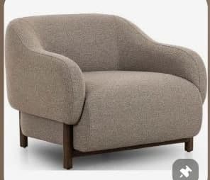 A contemporary accent chair featuring a compact and rounded silhouette. The chair is fully upholstered in a textured taupe or light brown woven fabric, covering the seat, back, and integrated arms. It stands on four slender, tapered dark brown wooden legs, which are visible beneath the upholstered base. The design emphasizes comfort and modern simplicity, with a soft, inviting form. — 3D model