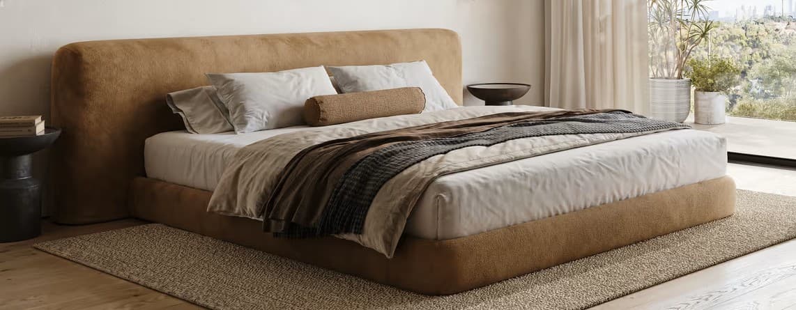 A modern platform bed featuring a fully upholstered frame and an integrated, slightly curved headboard. The bed has a low profile, giving it a minimalist and contemporary appearance, and is covered in a camel-colored, textured fabric. It is styled with white and neutral-toned bedding, including pillows and throws. — 3D model