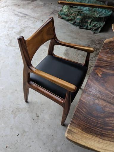 A Mid-Century Modern style dining armchair crafted from rich, dark wood with a distinct grain, featuring a contoured, integrated backrest and armrests, and an upholstered black leather seat. The chair's design emphasizes organic curves and sculpted lines. — 3D model
