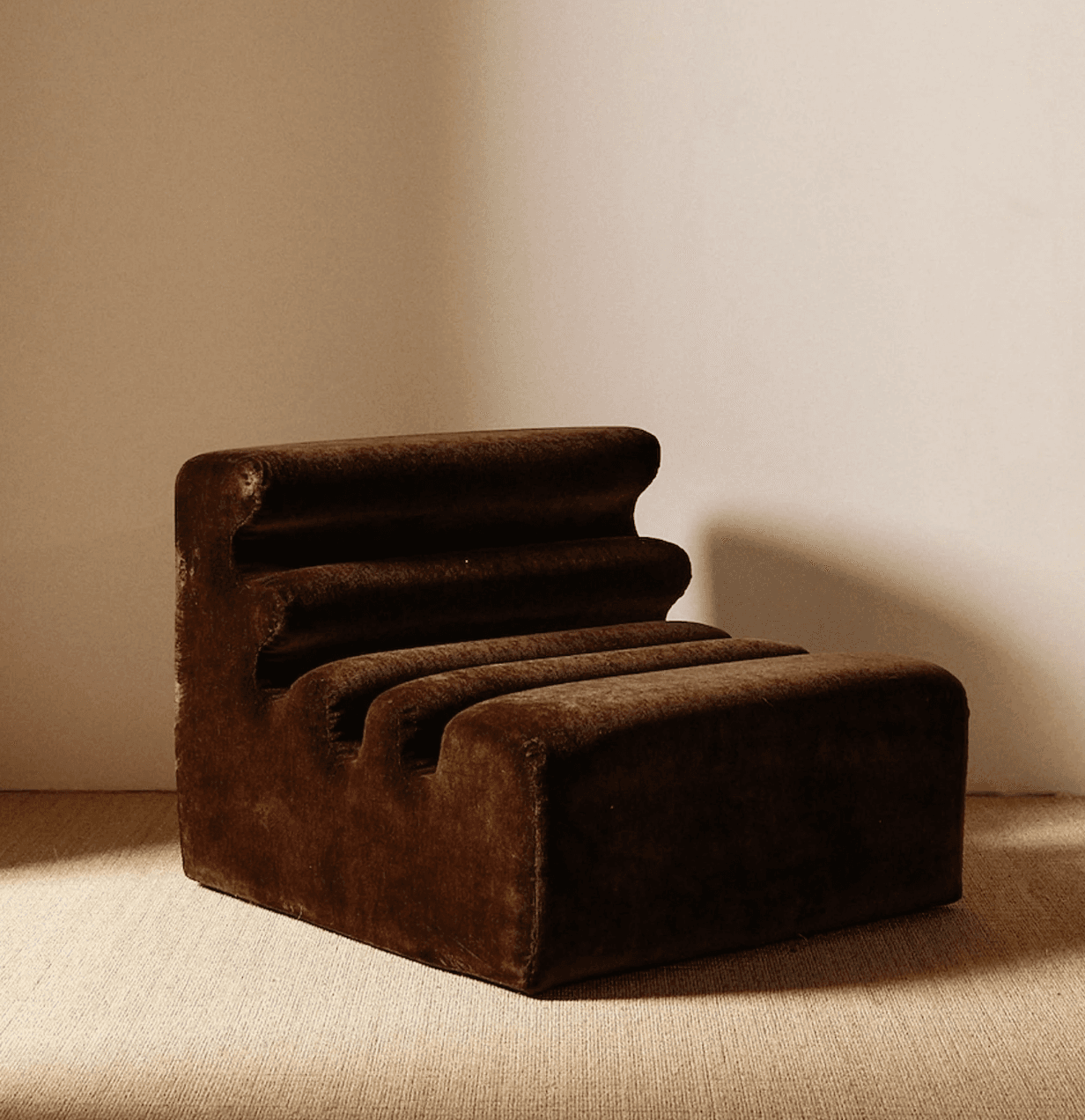 A low-profile, armless lounge chair upholstered in dark brown velvet or similar plush fabric, featuring a distinctive ribbed design on its backrest and seat. The chair has a sculptural, block-like form, with horizontal ridges creating a wave-like contour. It appears new and designed for comfort and aesthetic appeal in a contemporary setting. — 3D model