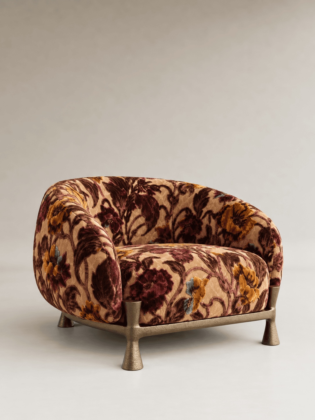 A compact, fully upholstered barrel-back lounge chair featuring a plush velvet fabric with an intricate floral pattern in shades of beige, burgundy, gold, and blue. The chair is supported by a distinctive, low-profile cast metal base with four sculptural legs in a bronze finish. — 3D model